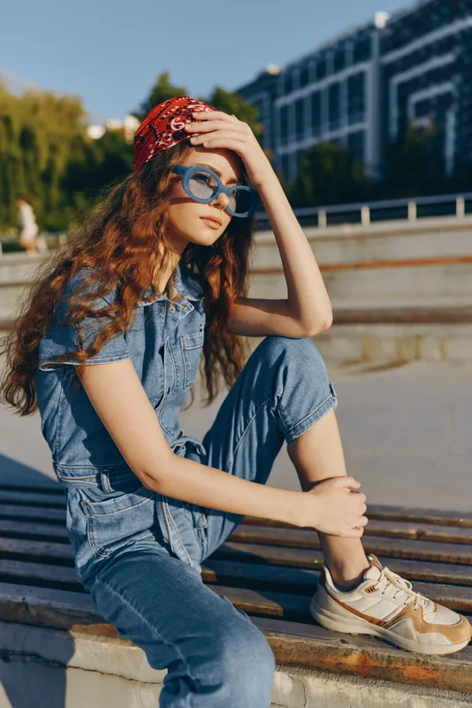 Lifestyle woman in modern boho-western denim street style wearing sunglasses and a red bandana, relaxing outdoors in warm natural light with film color effect.