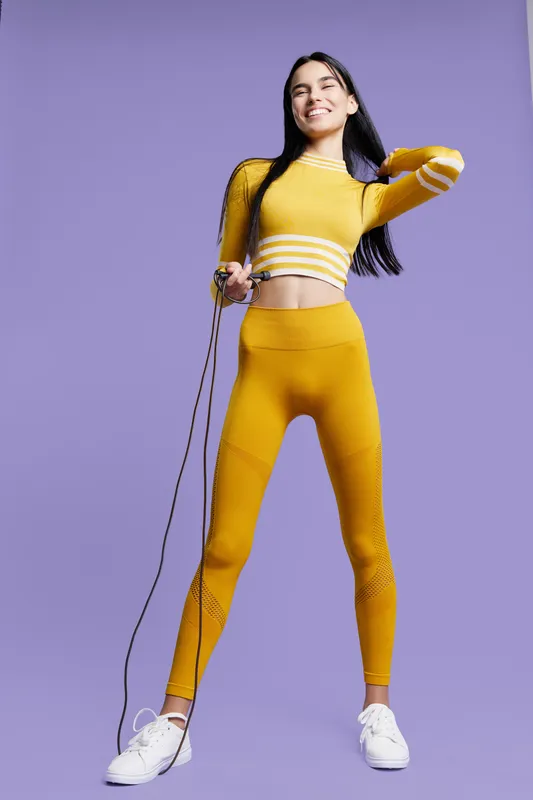 Young woman sport fitness jumping rope in bright yellow activewear with long sleeves, smiling and energetic, isolated on purple background for workout motivation.