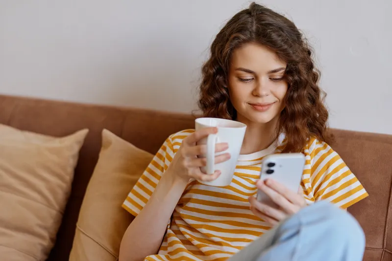 Young woman with wavy brown hair relaxes on a cozy sofa, holding a mug in one hand and scrolling on her phone, casual home setting. The scene conveys relaxation and everyday life. Bright.