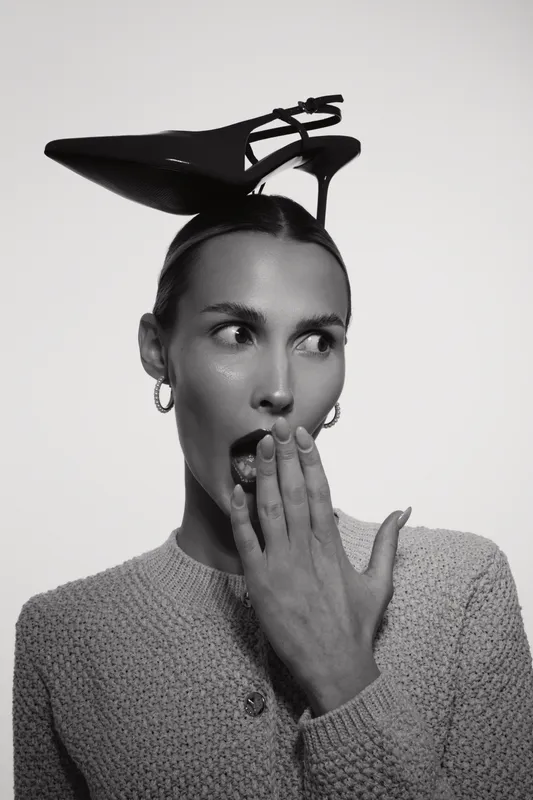 Surprised caucasian woman with light skin and dark hair holds hand near mouth, wearing high heel shoe on head in black and white studio portrait.