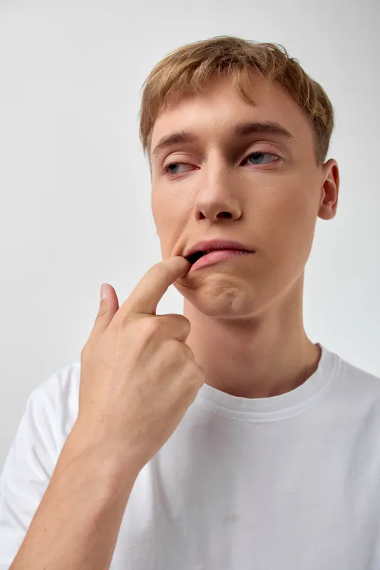 Thinking contemplative man with finger on lip, wearing a white shirt, conveying curiosity, hesitation and reflective mood in a simple studio setting