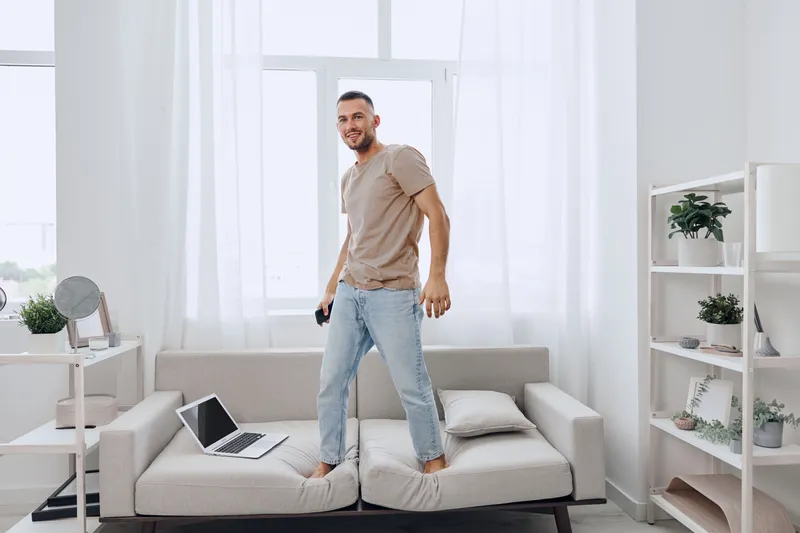 A smiling man stands on a couch, embodying the blend of work and life in a modern home. His relaxed demeanor reflects a digital detox amidst evolving mental health awareness.
