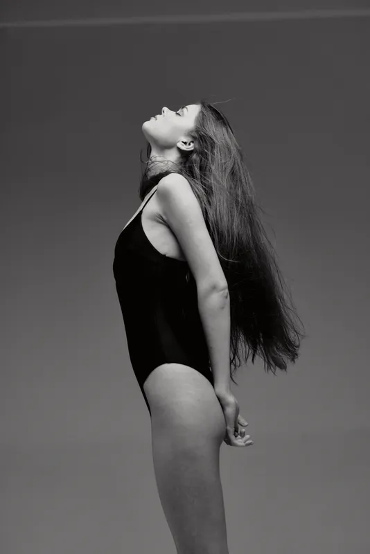 Black and white profile of a young woman with long hair wearing a black bodysuit, posing gracefully and looking upwards.