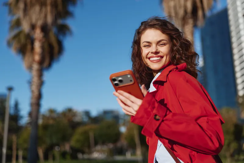 Young woman with curly hair smiles while holding a smartphone outdoors on a sunny day with palm trees in the background.