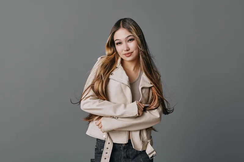 Young woman with long hair wearing beige jacket and black pants standing with arms crossed against grey background in studio portrait. Confident look and casual style on female model.