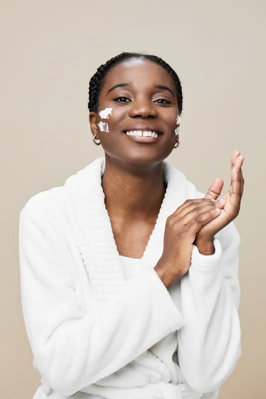 Smiling Black woman applying skincare cream in a cozy white robe, promoting beauty and self care