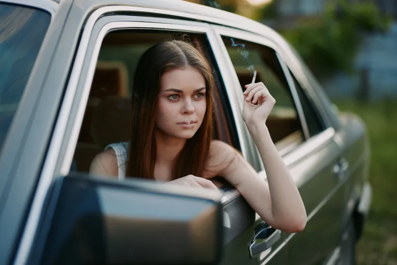 A thoughtful young woman sits in a vintage car, smoking a cigarette while looking out the window, capturing a moody summer vibe.