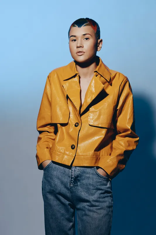 Young woman with short hair wearing a mustard yellow leather jacket and blue jeans stands confidently against a gradient blue background, showcasing modern fashion style.