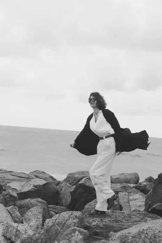 Woman wearing fashion forward attire stands on rugged rocks along the coastline by the sea, wind catching flowing jacket, conveying movement and elegance in a monochrome outdoor scene
