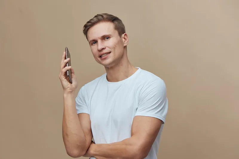 Smiling young man in casual white t-shirt holding mobile phone close to ear, looking friendly, standing against plain beige background, studio portrait. People lifestyle concept.