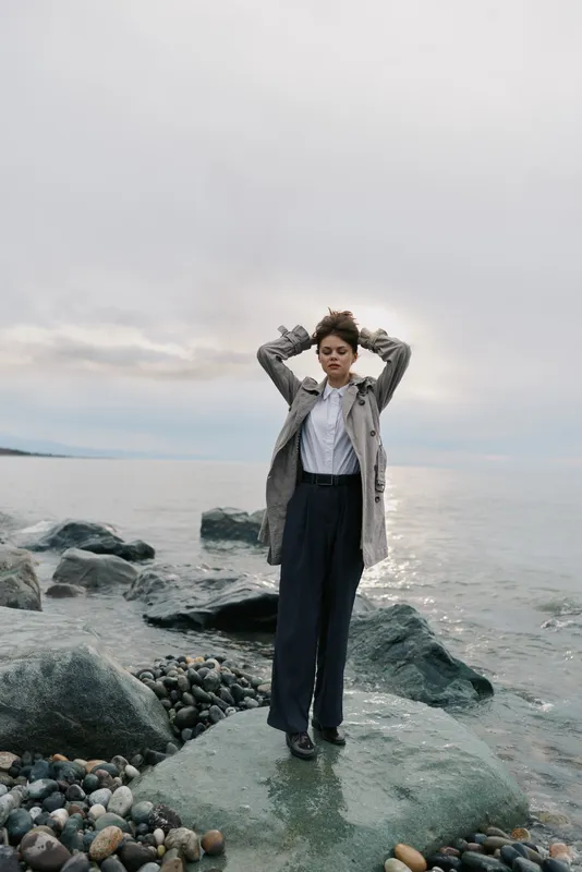Young woman in casual clothes stands on rock by sea with eyes closed and hands behind head. Overcast sky and calm water create peaceful, contemplative mood outdoors.