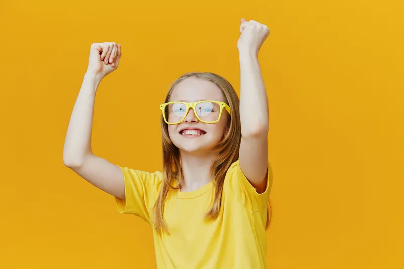 Cheerful young girl celebrating success with raised fists, wearing yellow glasses and a bright yellow shirt against a vibrant yellow background