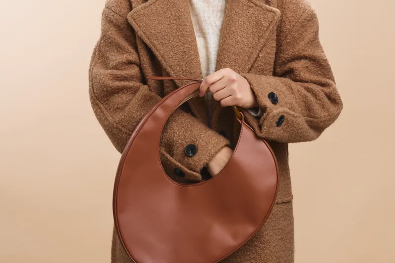 asian woman wearing a textured brown coat holds a circular leather bag against a neutral backdrop. the scene conveys chic, understated style and a calm, fashionable mood.