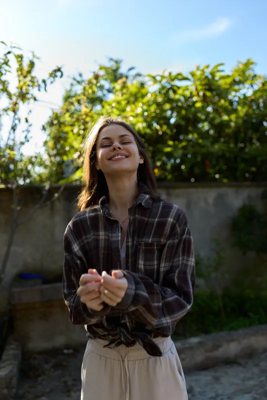 Young woman wearing a plaid shirt stands outdoors, smiling softly under warm light with green foliage in the background, a relaxed casual moment captured in daylight