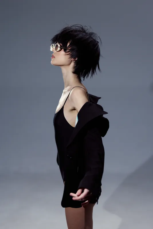 Stylish woman in black dress wearing sunglasses with short hair posing on gray background