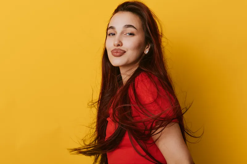 Young woman with long hair in red shirt posing on yellow background with confident expression and slight smile. Studio portrait with vibrant colors and casual style.