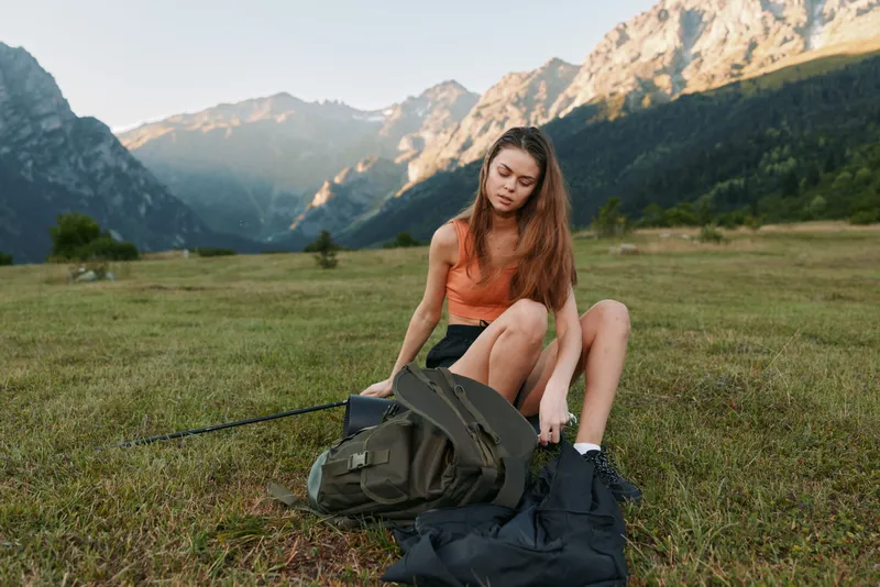 A young woman sits on a grassy meadow with a backpack, in a rugged mountain landscape. She adjusts gear, enjoying a calm, adventurous outdoor moment.