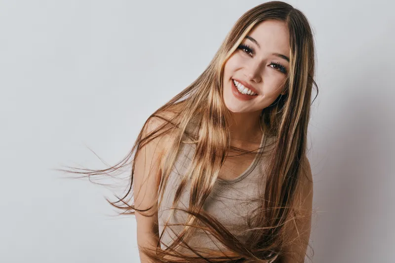 Smiling woman with long hair wearing casual sleeveless top posing against white background. Portrait of happy girl with natural makeup and flowing hair indoors in bright studio setting.