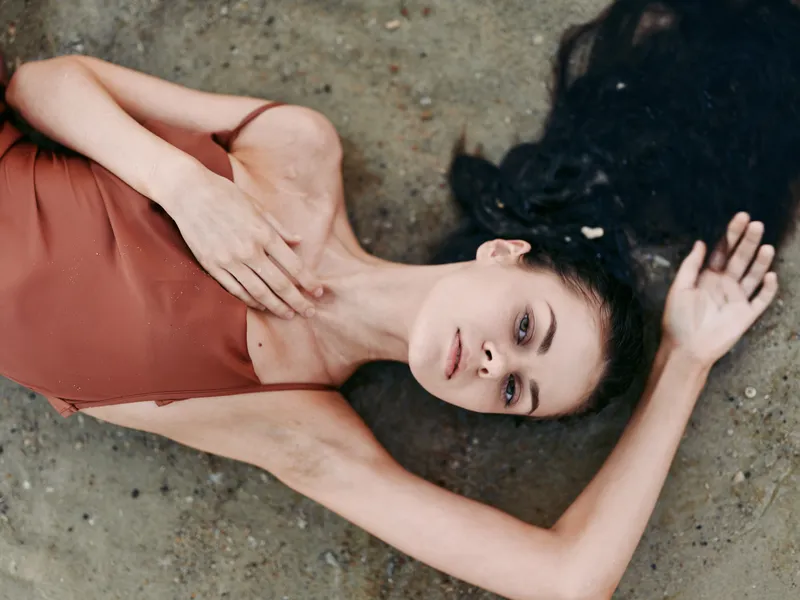 A woman lies on a textured ground wearing a rust colored dress, head turned, arms extended, creating a calm, intimate pose with soft lighting and a moody atmosphere.