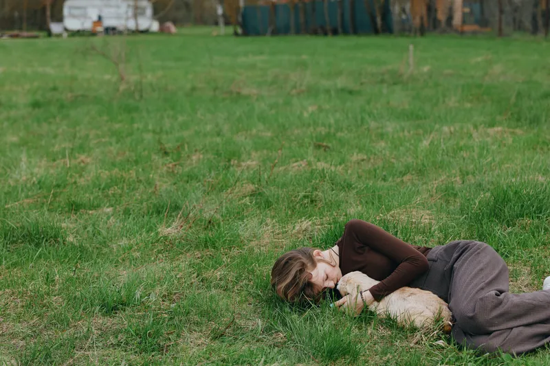 Woman lying on grass cuddling a goat kid outdoors, showing countryside relaxation and gentle animal companionship. Warm natural light creates a calm, peaceful mood and sense of connection with nature.