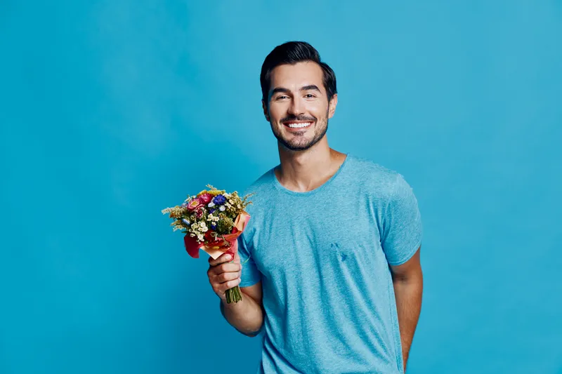 Happy smiling young man holding a bouquet of flowers, wearing a blue t-shirt, standing against a vibrant blue background. Bright cheerful lifestyle portrait.