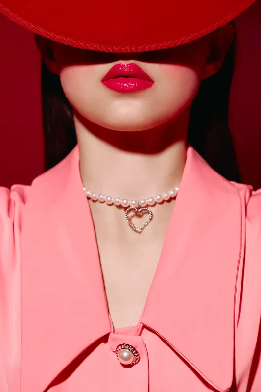 Portrait of woman with bright red lipstick wearing pearl necklace with heart pendant and pink suit, partially covered by large red hat. Concept of beauty, fashion, elegance, and luxury.