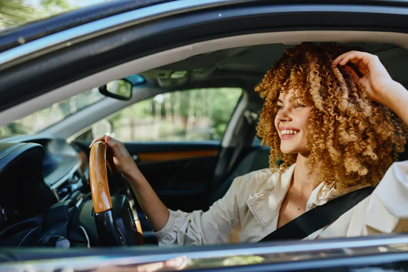 Smiling woman driving a car with curly hair wearing a beige jacket and seatbelt, enjoying a sunny day in the suburban area with green trees visible outside.