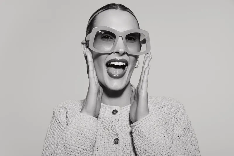 Happy Caucasian woman with light skin wearing large glasses and knitted sweater smiles with open mouth isolated on grey background.