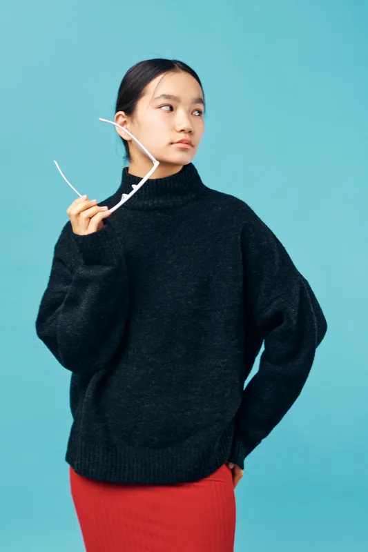 Young woman in black oversized sweater holding white eyeglasses with one hand wearing red skirt on blue background looking sideways. Casual fashion, thoughtful pose and modern style portrait.