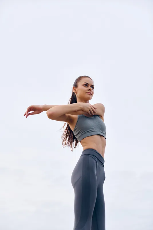 Young athletic woman stretching outdoors in stylish sportswear against a soft grey sky, embodying fitness and health concepts, perfect for wellness and active lifestyle themes