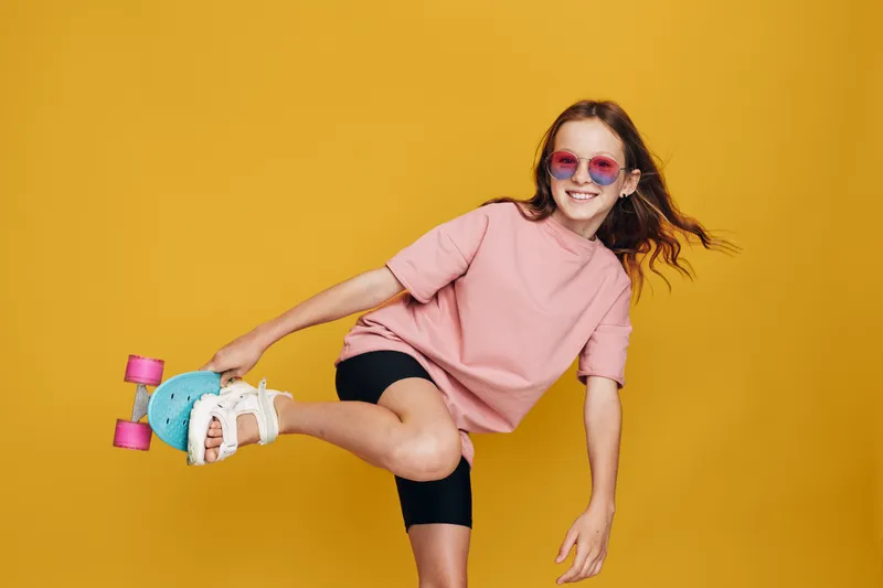 Happy girl in pink sunglasses and t-shirt roller skating on one leg against yellow background. Youthful energy and fun concept with motion and joy in studio lighting.