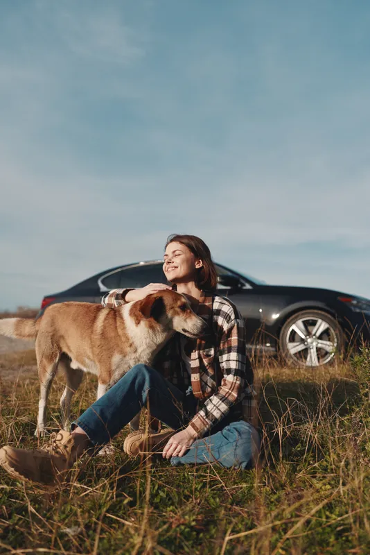Woman sits in a grassy field with a dog beside her, car in the background, enjoying a quiet moment in nature, casual clothing, warm sunlight, peaceful companionship and rural scenery