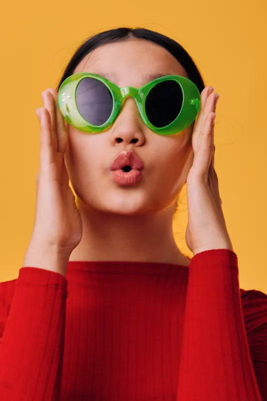 Young woman with green sunglasses making surprised facial expression posing on orange background. Fashion portrait of female in red top with modern eyewear and playful mood in studio.
