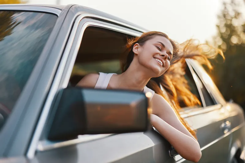 A young woman enjoying the summer breeze while leaning out of a car window, exuding happiness and freedom with her hair flowing in the wind.