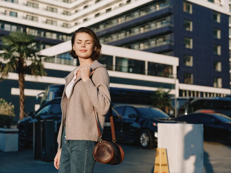 A cheerful woman stands in a modern outdoor setting beside a glass building. She smiles warmly, carrying a small leather bag, exuding confidence and relaxed style.