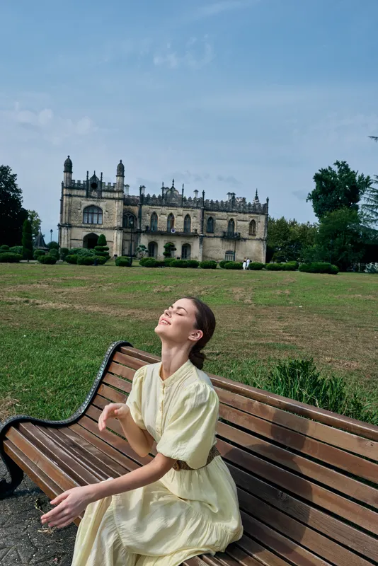 Woman on bench in park near historic building wearing yellow dress enjoying sunlight relaxation and happiness authentic inclusive portrait with calm smile