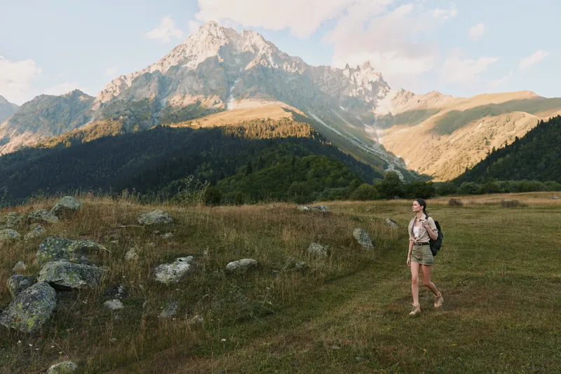 Young woman hiking through a sunlit mountain meadow with a backpack, enjoying open space, fresh air, and a sense of freedom.