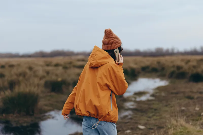 Woman outdoors in countryside, wearing a yellow jacket and orange beanie, walking beside a stream, holding a phone to her ear, winter lifestyle, thoughtful pose, windy air, soft light