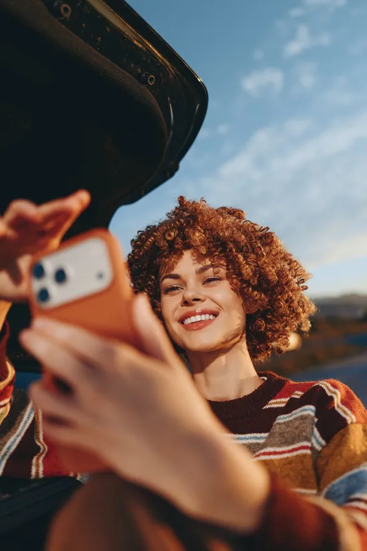 Woman lifestyle selfie outdoors, curly hair smiling in a travel landscape, she holds a smartphone for casual photo, warm sunlight, friendly expression, close up portrait today