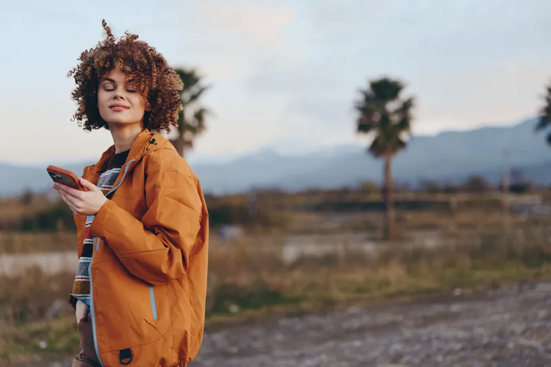 Woman lifestyle portrait, curly hair woman in an orange jacket, standing outdoors on a rural path, looking at a phone, calm travel mood, mountains and trees softly blurred, natural daylight