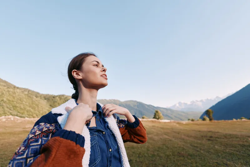 Woman enjoying nature in mountains and meadow, outdoor portrait of a young traveler hiking with jacket and backpack, travel lifestyle scene showing freedom, sunlight and peaceful horizon.