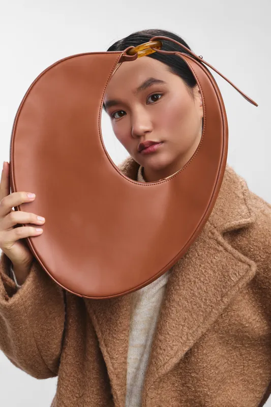 asian woman wearing a warm coat holds a large circular bag to her face, creating a stylish and playful portrait in a clean studio setting.