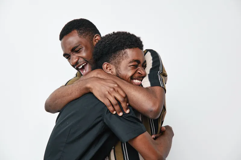 Two young black men with dark skin laughing and hugging each other in casual clothes on white background. Friendship, happiness and joyful moment between male friends.
