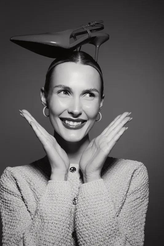 Smiling white woman with fair skin and earrings balances a high heel shoe on her head, posing playfully with hands near face in studio lighting.
