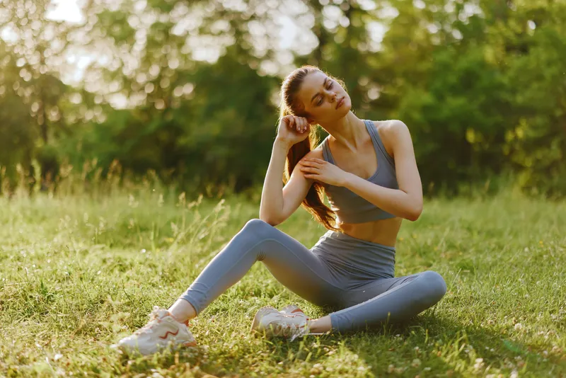 Athletic young woman sitting in a serene park during sunset, wearing comfortable sportswear and expressing relaxation, perfect for wellness and fitness concepts