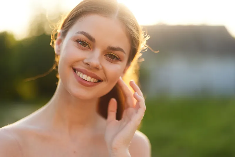 Young smiling woman gently touching her hair in a sunlit outdoor setting, showcasing natural beauty and warmth perfect for health and lifestyle concepts