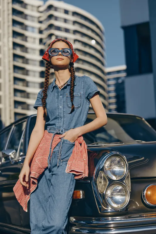 stylish young woman with braids wearing sunglasses and casual denim outfit leaning on vintage car in urban sunny setting with modern buildings in background