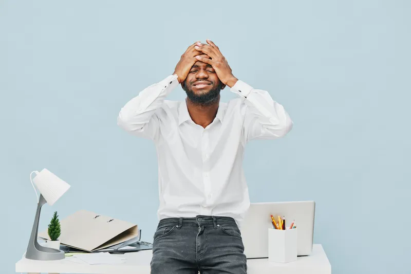 Frustrated young African American man with beard, wearing white shirt, holding head with hands, standing in modern office with desk, laptop, and stationery, expressing stress or anxiety, studio shot, 