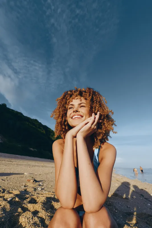Smiling woman on beach with curly hair, sunlight on skin, sitting outdoors and touching her face, concept of skincare self care and wellness in summer, warm mood relaxation and peace