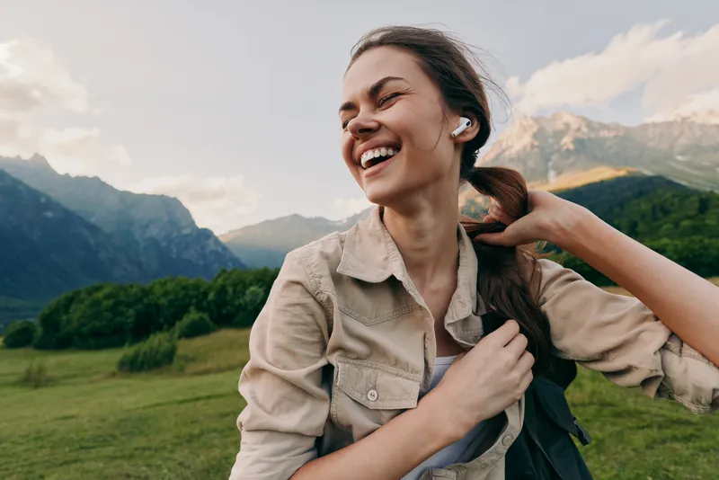 A joyful woman outdoors, wearing a light jacket, backpack over her shoulder, smiling as green fields and distant mountains frame the scene. Lifestyle moment of freedom and nature.
